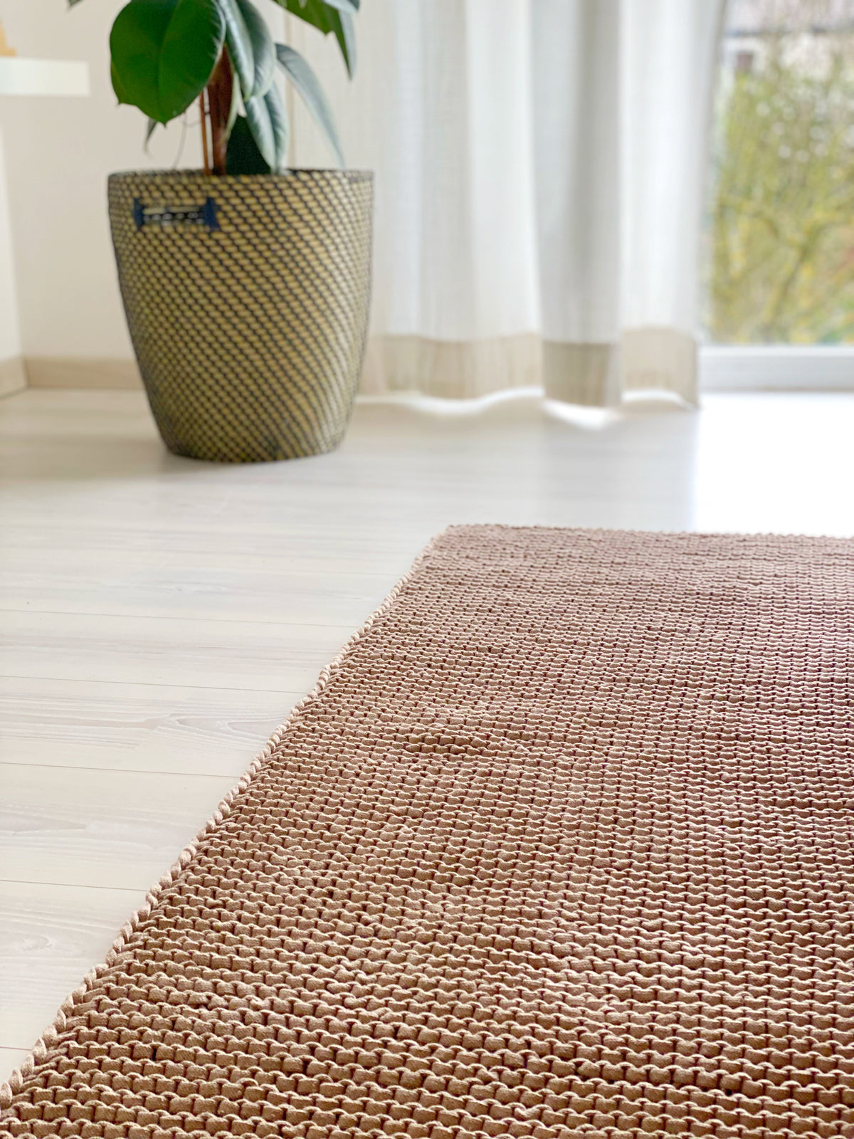 Leinenfarbener Teppich aus groben Recyclinggarn auf einem weißen Boden. Im Hintergrund eine Pfanze in einem Bastkorb. Beispiel für eine individuelle Anfertigung von Stadtrandstil handmade