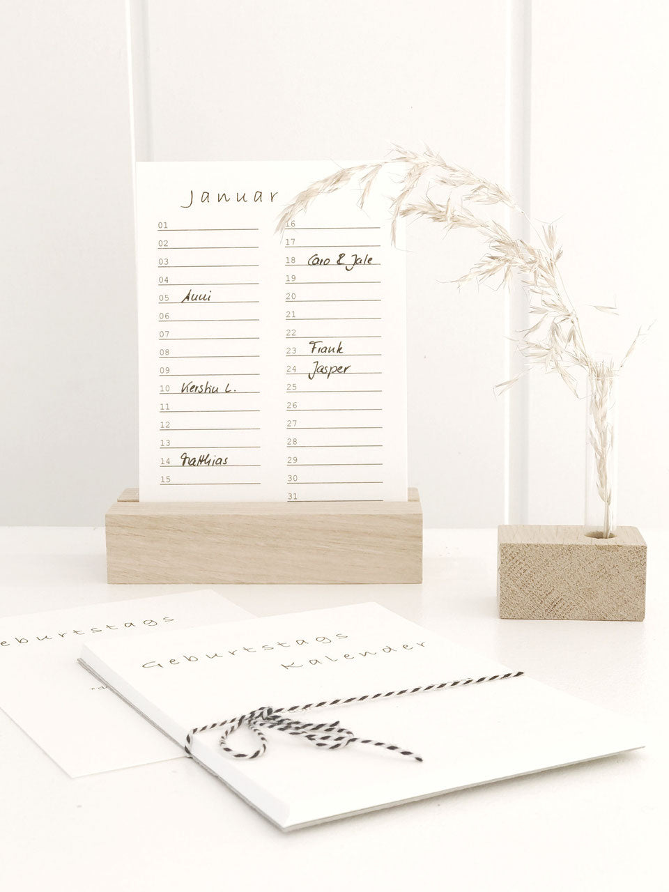 Geburtstagskalender (immerwährend) in minimalistischem Design von Stadtrandstil in Postkartenformat mit einem Halter aus unbehandeltem Eichenholz. Daneben eine Minivase mit getrocknetem Gras.
