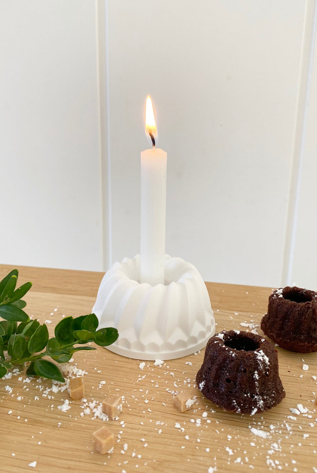 Ein Kerzenständer in Gugl Form mit weißer, brennender Kerze. Als Geburtstagsidee mit kleinen Schokoladen Gugln, bestreut mit Puderzucker. Daneben ein kleiner Buchsbaumzweig.