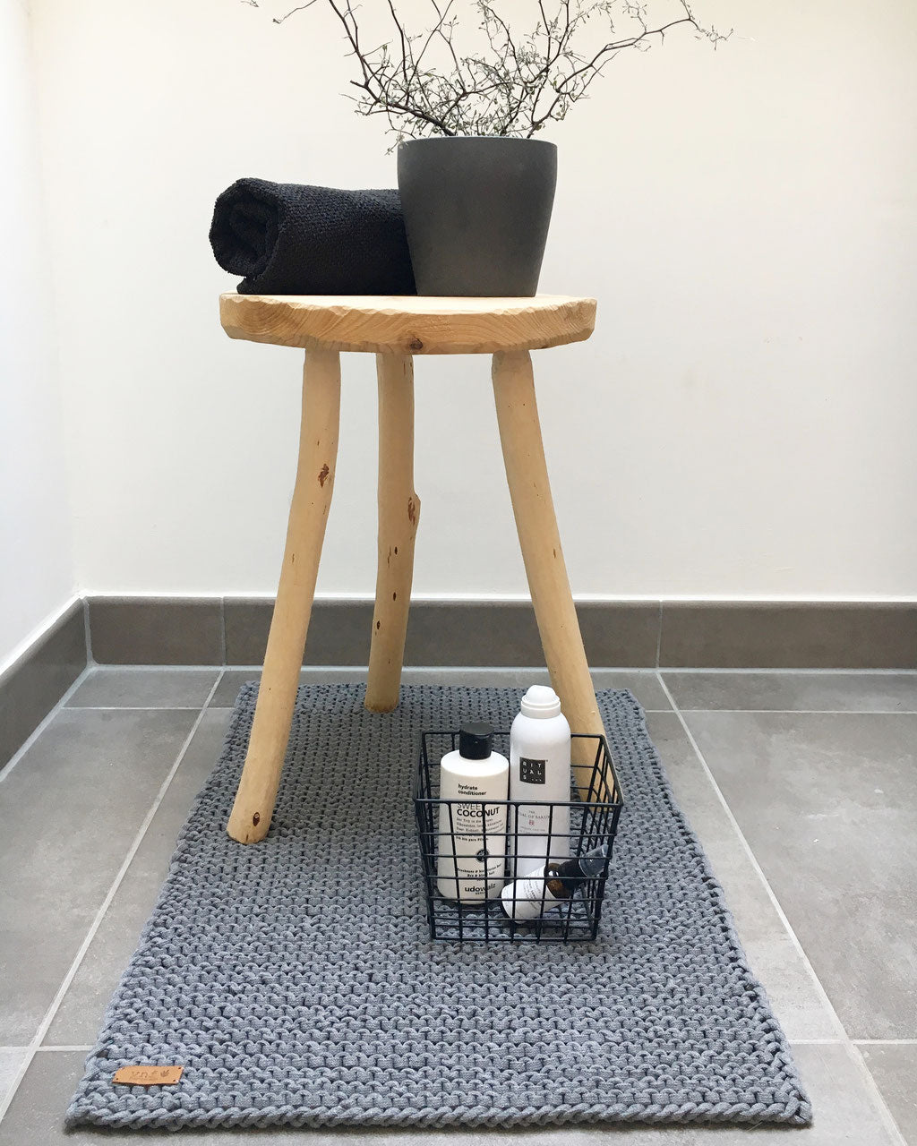 Dunkelgrauer Badvorleger aus Recyclinggarn. Dekoriert mit Badezimmer Utensilien : einem Drahtkorb mit Pfelgeprodukten und einem Holzhocker mit schwarzem Badezimmer natürlich.andtuch und einer Pflanze. Badinspiraion Stadtrandstil