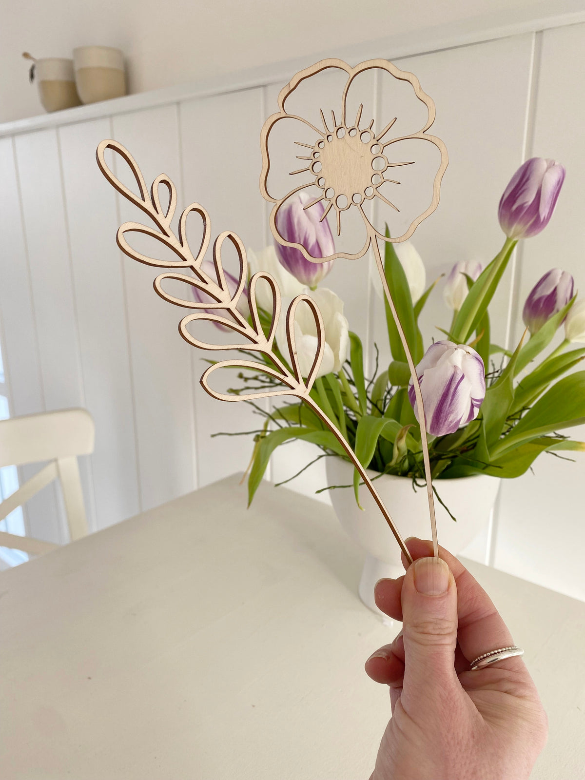 zwei lasercut Holzblumen in der Hand. Einmal wine minimalistische Blume, einmal ein Gras. Dahinter ein weißer Küchentisch mit einer Schale mit lila Tulpen und zwei Bechern