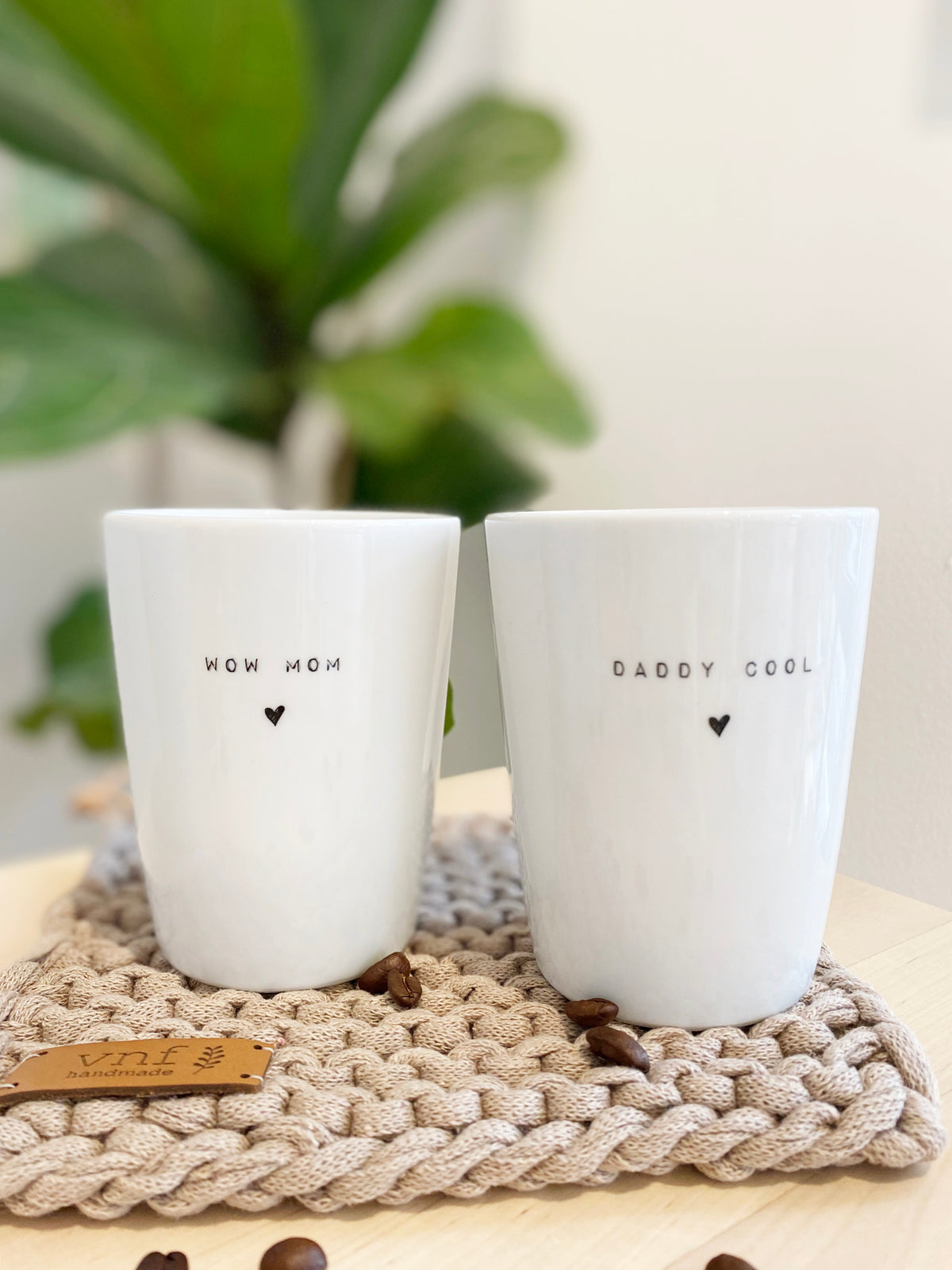 zwei weoße Becher aus Porzellan mit dem Aufdruck "wow mom" und "daddy cool". Natürlich arrangiert auf handgestrickten Untersetzer mit Grünpflanze und Kaffeebohnen