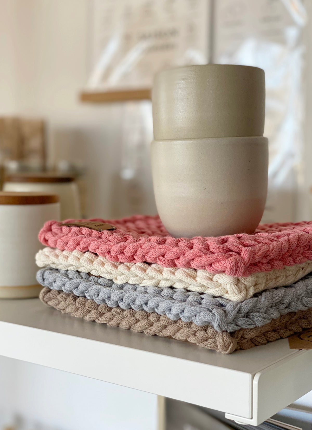 Regal mit Küchenartikeln bei Stadtrandstil. Stapel mit vier Untersetzern. hand gestrickt in unterschiedlichen Naturtönen und zwei Tonbechern