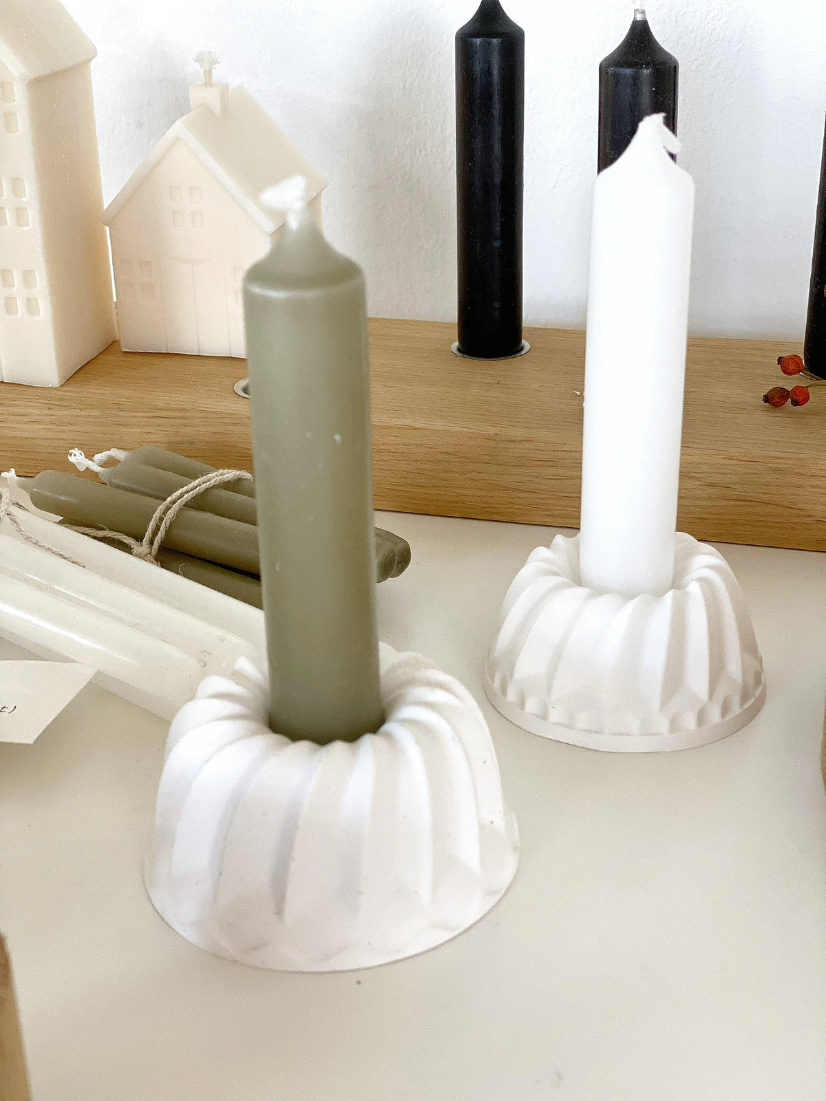 zwei weiße Kerzenhalter aus Raysin in Guglform mit Kerzen, einmal oliv, einmal weiß. Dahinter liegen weitere Kerzen und ein Kerzenbrett aus Eiche, dass ebenfalls mit Kerzen dekoriert ist-