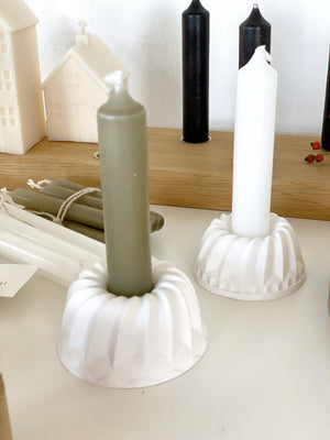 zwei weiße Kerzenhalter aus Raysin in Guglform mit Kerzen, einmal oliv, einmal weiß. Dahinter liegen weitere Kerzen und ein Kerzenbrett aus Eiche, dass ebenfalls mit Kerzen dekoriert ist-