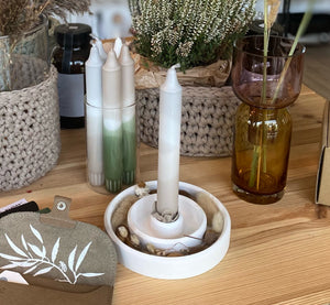 Weiße Kerzenschale mit doppeltem Rand, gefüllt mit Naturmaterialien und weißer Kerze. Zusammen auf einem Holztosch mit schemenhaft erkennbaren Häkelkörben, einer Vase, Kerzen und einer kleinen Ledertasche. Herbstliche Impression von Stadtrandstil