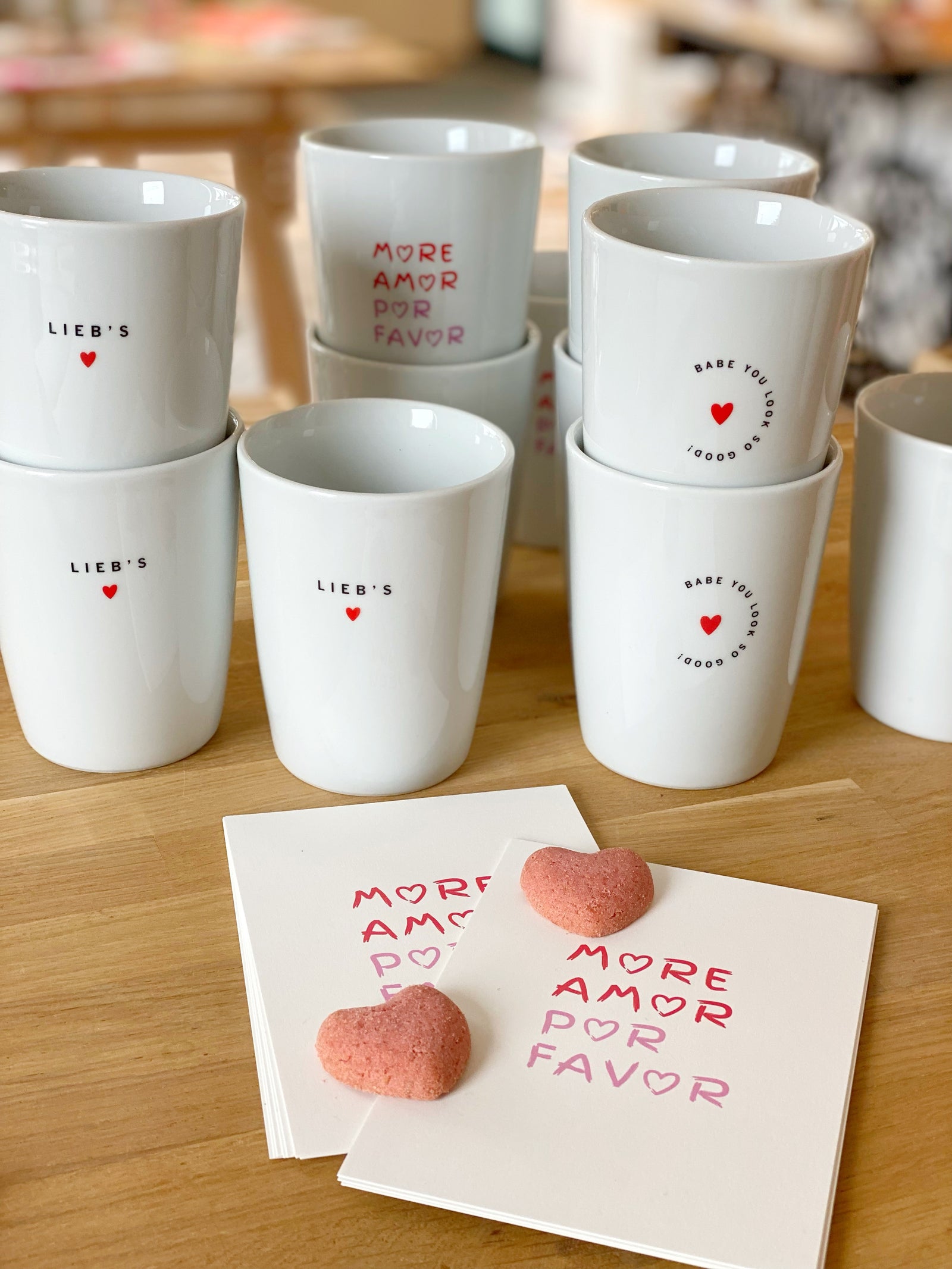 Mehrere ineinandergestapelte, weiße Kaffeebecher mit Aufschrift "lieb's" , und Herzen davor Postkarten mit dem Aufdrck "more amor por favor" und zwei Keksen in Herzform