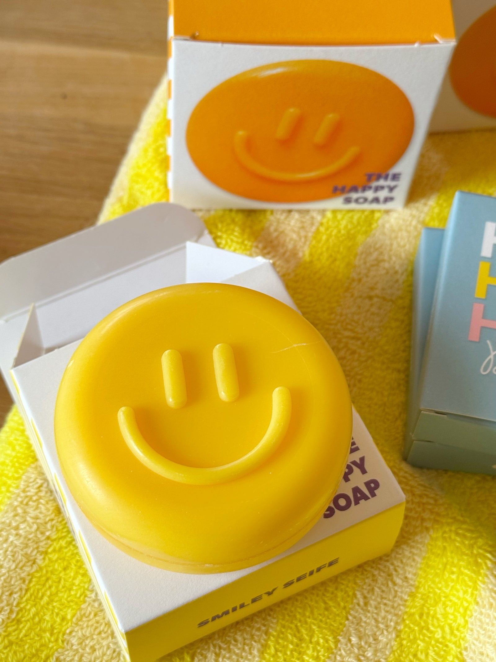 gelbe, runde seife mit Smiley auf dazugehöriger Verpackung, im Hintergrund die Seife in orange