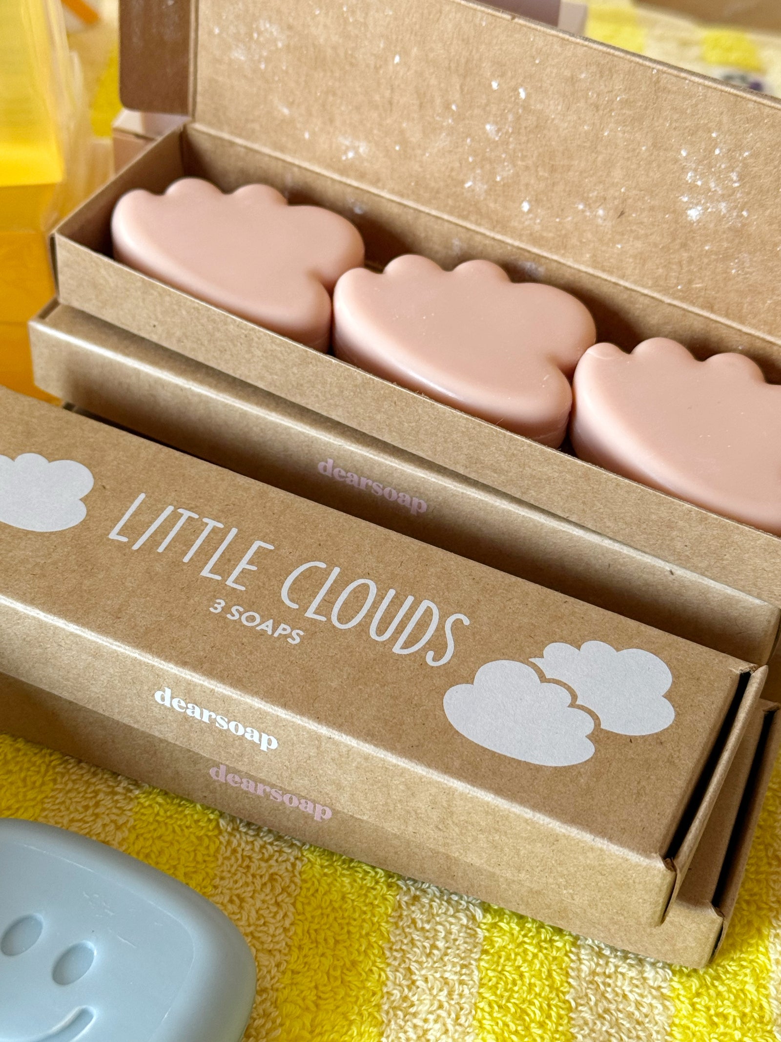 drei kleine rosafarbene Seifen in Wolkenform in geöffneter Verpackung aus Kraftpapier, davor eine geschlossene Verpackung