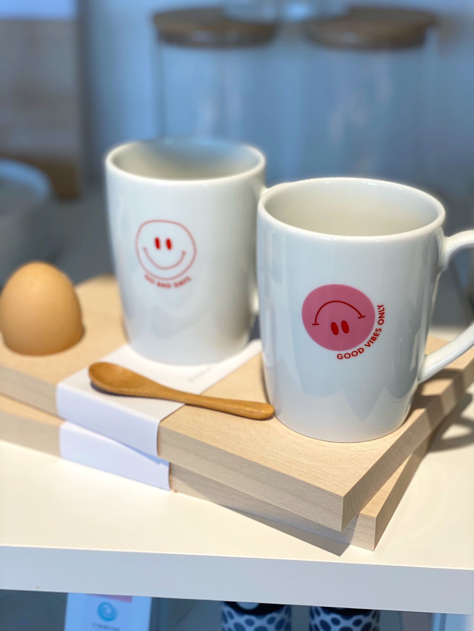 Zwei weiße Porzellantassen mit Smiley Aufdruck auf Frühstcksbrett mit Löffel und Ei