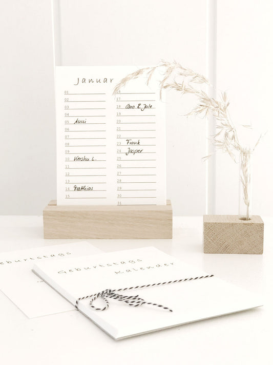 Geburtstagskalender (immerwährend) in minimalistischem Design von Stadtrandstil in Postkartenformat mit einem Halter aus unbehandeltem Eichenholz. Daneben eine Minivase mit getrocknetem Gras.