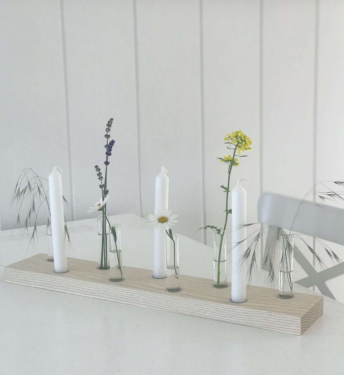 Großes Kerzen- und Blumenbrett mit Platz für neun Kerzen oder Reagenzgläsern. Dekoriert mit drei weißen Kerzen und Sommerblumen. Das Brett steht auf einem weißen Tisch vor einer weißen Holzwand