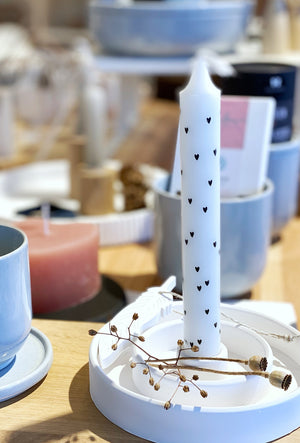 Weißer Kerzenständer, alch mit doppeltem Rand und einer weißen Kerze mit schwarzen Herzen sowie getrockneten Mohnkapseln