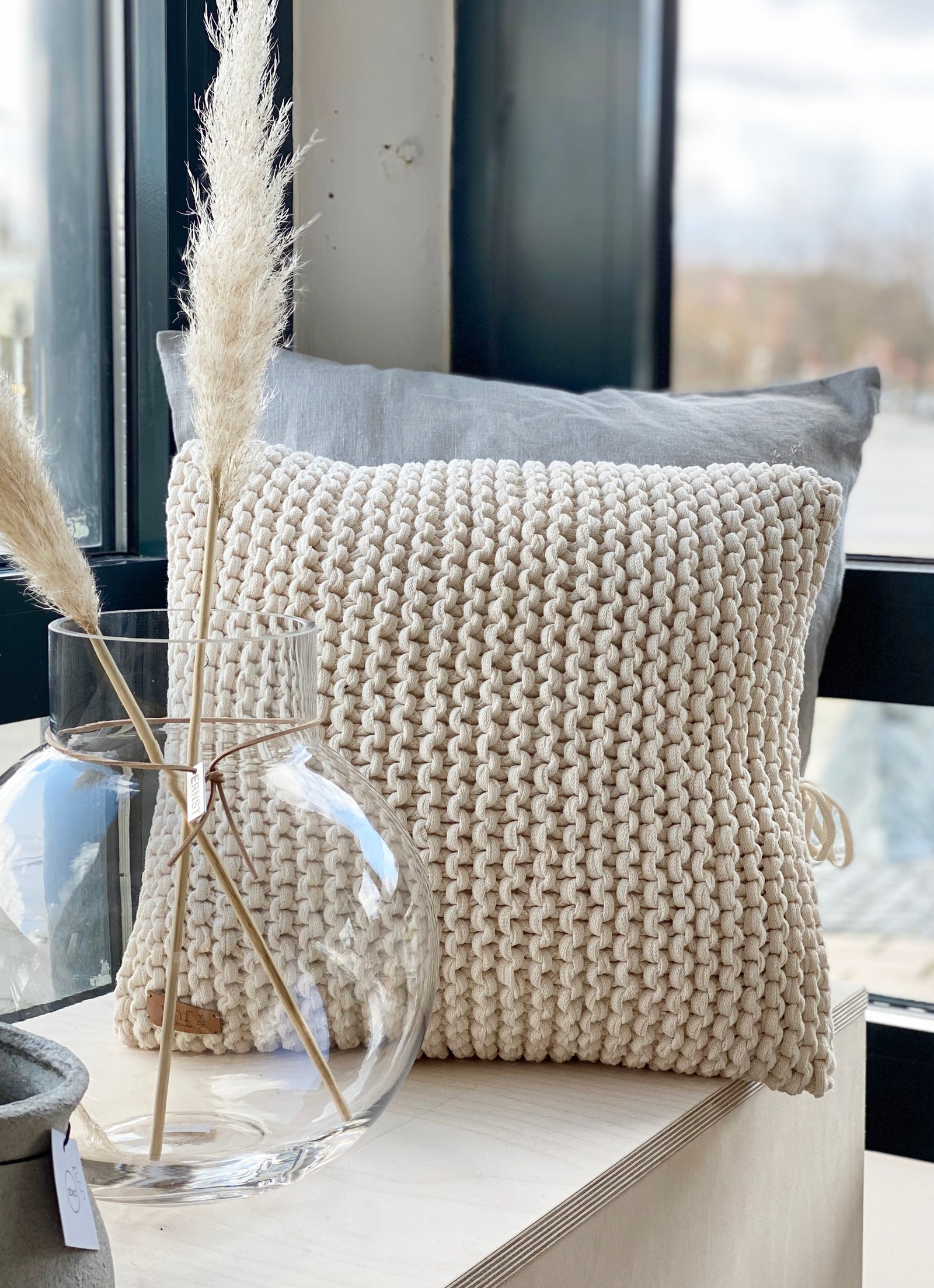 Naturfarbenes Kissen grob gestrickt auf einer Holzbank. Davor eine bauchige Glasvase mit getrockneten Gräsern. Eine natürliche Dekoration von Stadtrandstil