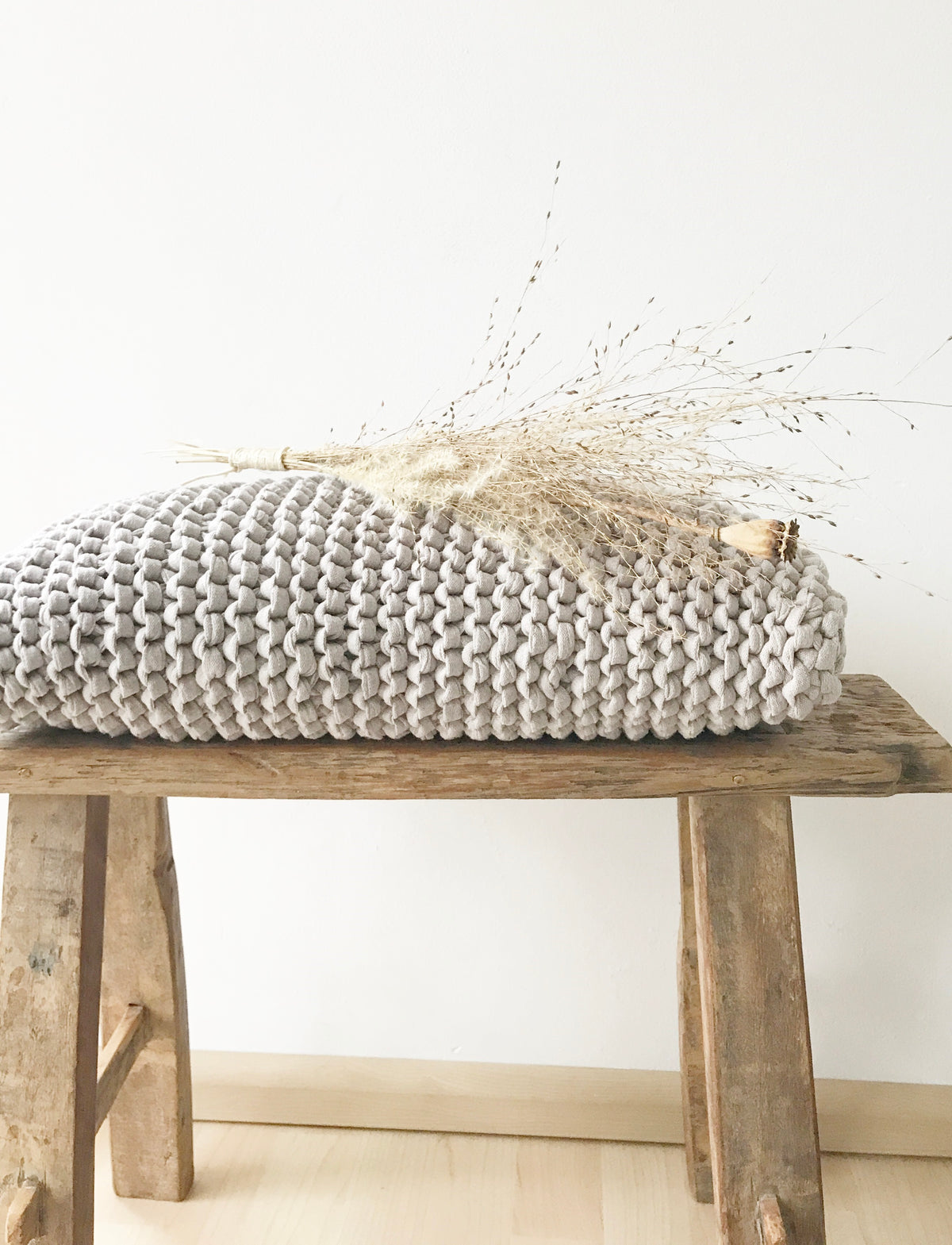 Hellgraues, handgestricktes Kissen aus Recyclingmix von Stadtrandstil. Natürlich dekoriert auf einer kleinen Holzbank aus Altholz und einem Bund getrockneter Gräser