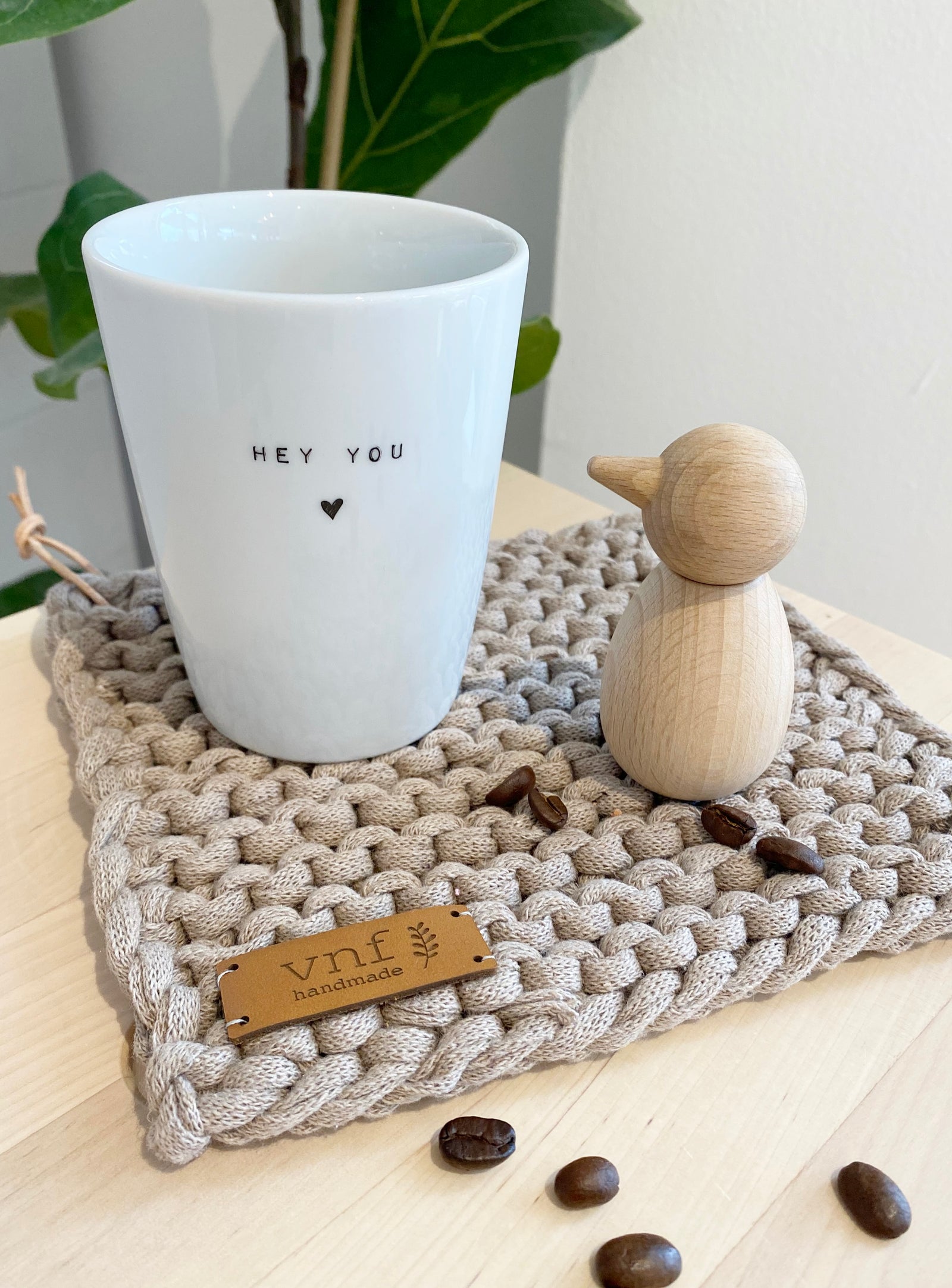 Hübsch arrangiertes Küchenbild: ein grauer, handestrickter Untersetzer, auf dem en kleiner Holzvogel sowie ein weißer Becher mit der AUfschrift "hey you" stehen. kaffeebohnen sind darüber getreut