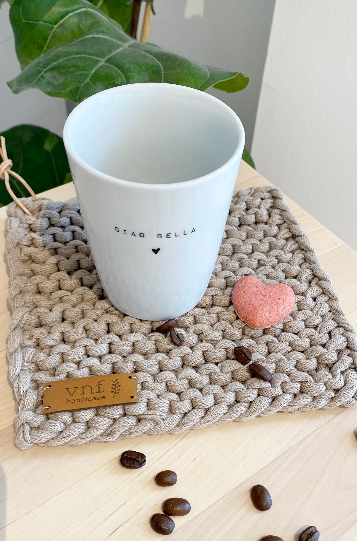 Natürlich dekorierer weißer Porzellan Becher mit dem Aufdruck "ciao bella" auf einem leinenfarbenen Untersetzer mit Lederlabel, dekoriert mit einem Keks in Herzform und Kaffeebohnen
