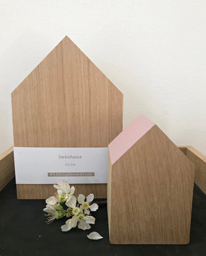 Großes, naturbelassenes Dekohaus aus Eiche zusammen mit einem kleinen, desssen Giebel rosé lackiert st. MIt zarter, weißer Blume arrangiert