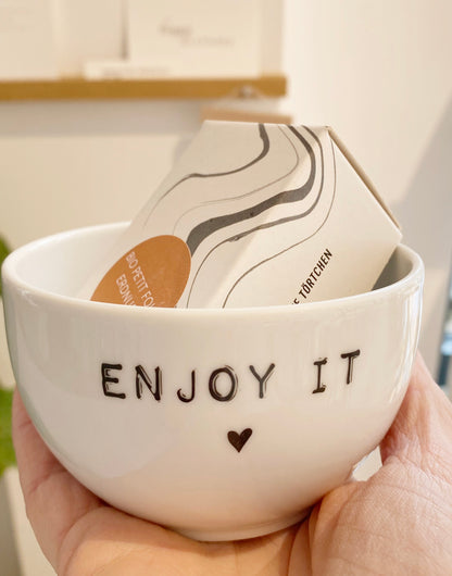 EIne weiße Dipschale mit den Aufdruck "enjoy it", gefüllt mit ener kleinen Törtvhen Schachtel wird in der Hand gehalten