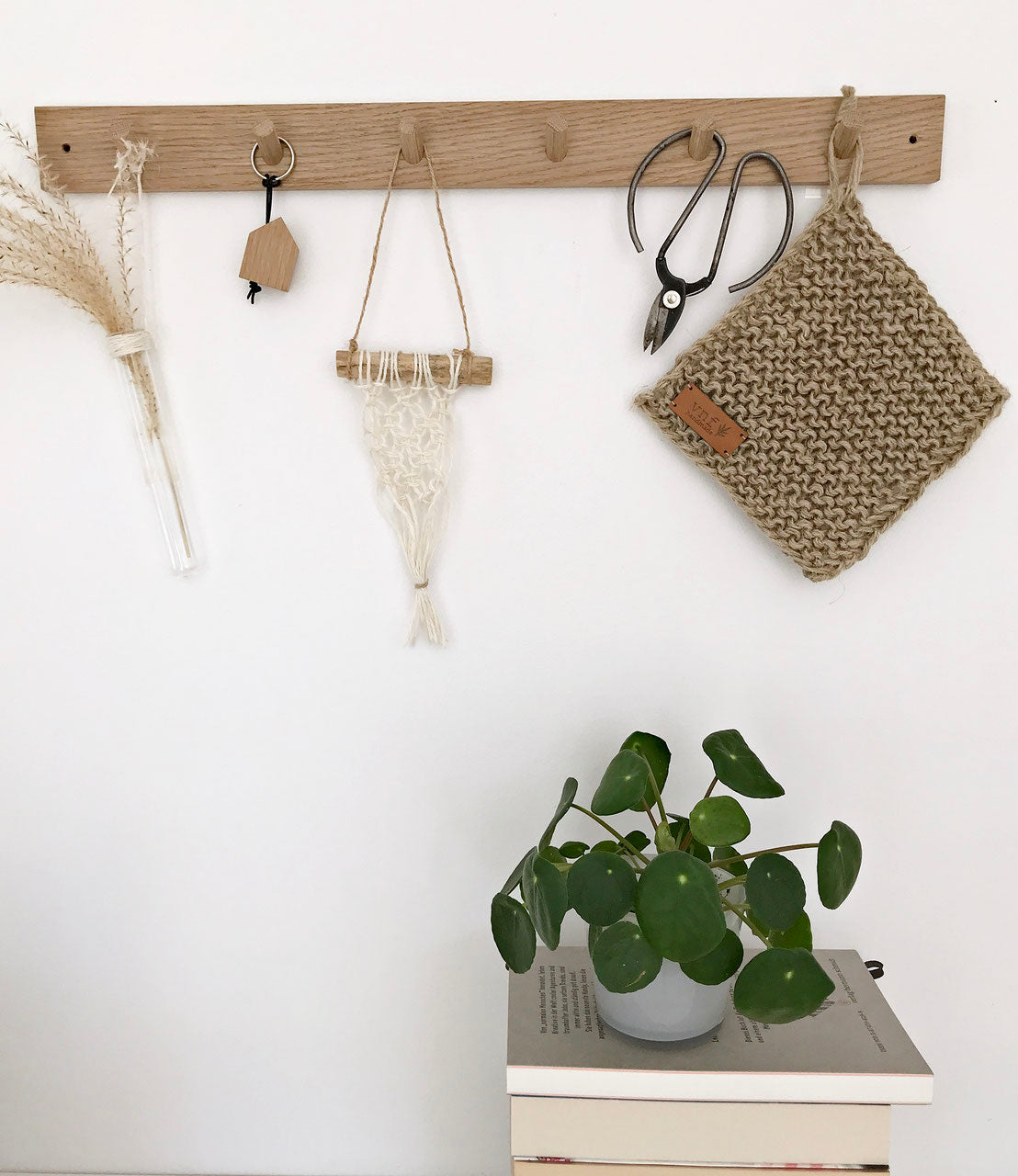 Große Hakenleiste von Stadtrandstil aus Eiche. Dekoriert mit Trockengräsern, einem Untersetzer aus Jute, einem kleinen Makramee, einer Schere und einem Schlüsselanhänger in Hausform. Darunter ein paar Bücher und eine Pilea Pflanze