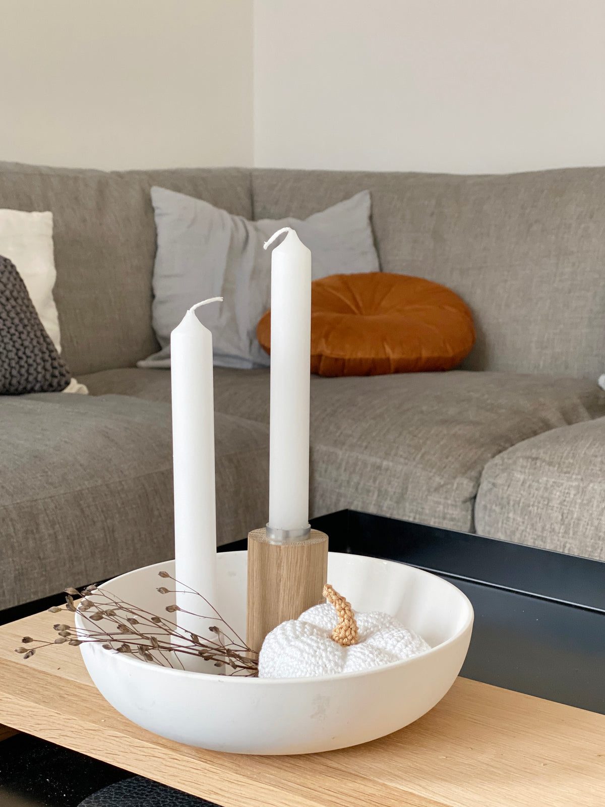 Wohninspiration : vor einem grauen Sofa mit Kissen steht ein schwarzer Tisch mit einem Holztablett. Darauf steht eine weiße Keramikschale, gefüllt mit getrockneten Pflanzen, einem Kerzenständer aus Eiche mit weißer Kerze und einem gehäkelten Kürbis. Eine natürliche Wohnzimmer Dekoration