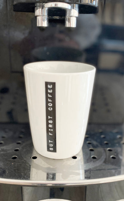 Weißer Porzellan Kaffeebecher mit schwarzem Aufdruck "but first coffee" unter Kaffeeausfluss einer Kaffeemaschine