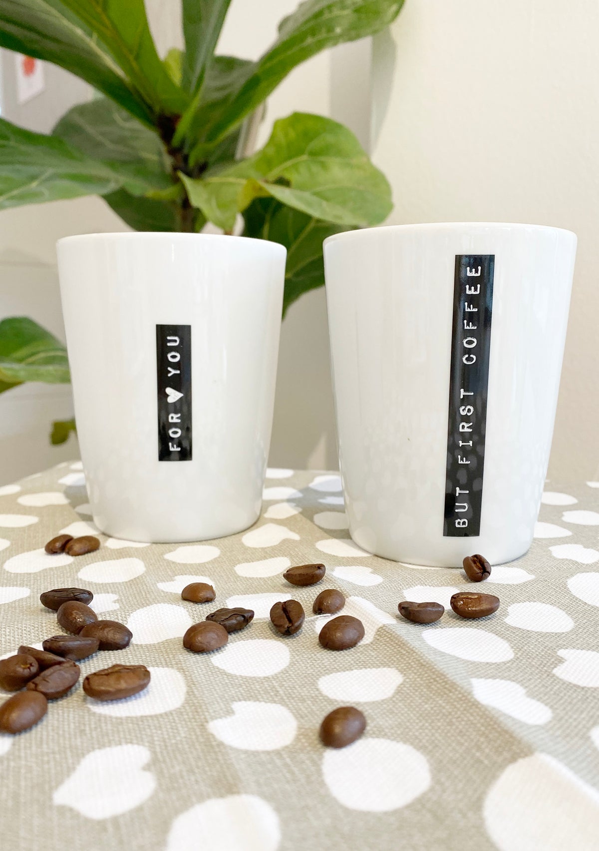 Zwei natürlich dekorierte Kaffeebecher mit dem schwarzen Aufdruck " for you" und "but first coffee", mit beige weißem Tuch, Kaffeebohnen und Grünpflanze