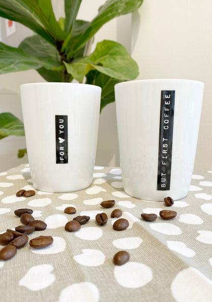 Zwei natürlich dekorierte Kaffeebecher mit dem schwarzen Aufdruck " for you" und "but first coffee", mit beige weißem Tuch, Kaffeebohnen und Grünpflanze