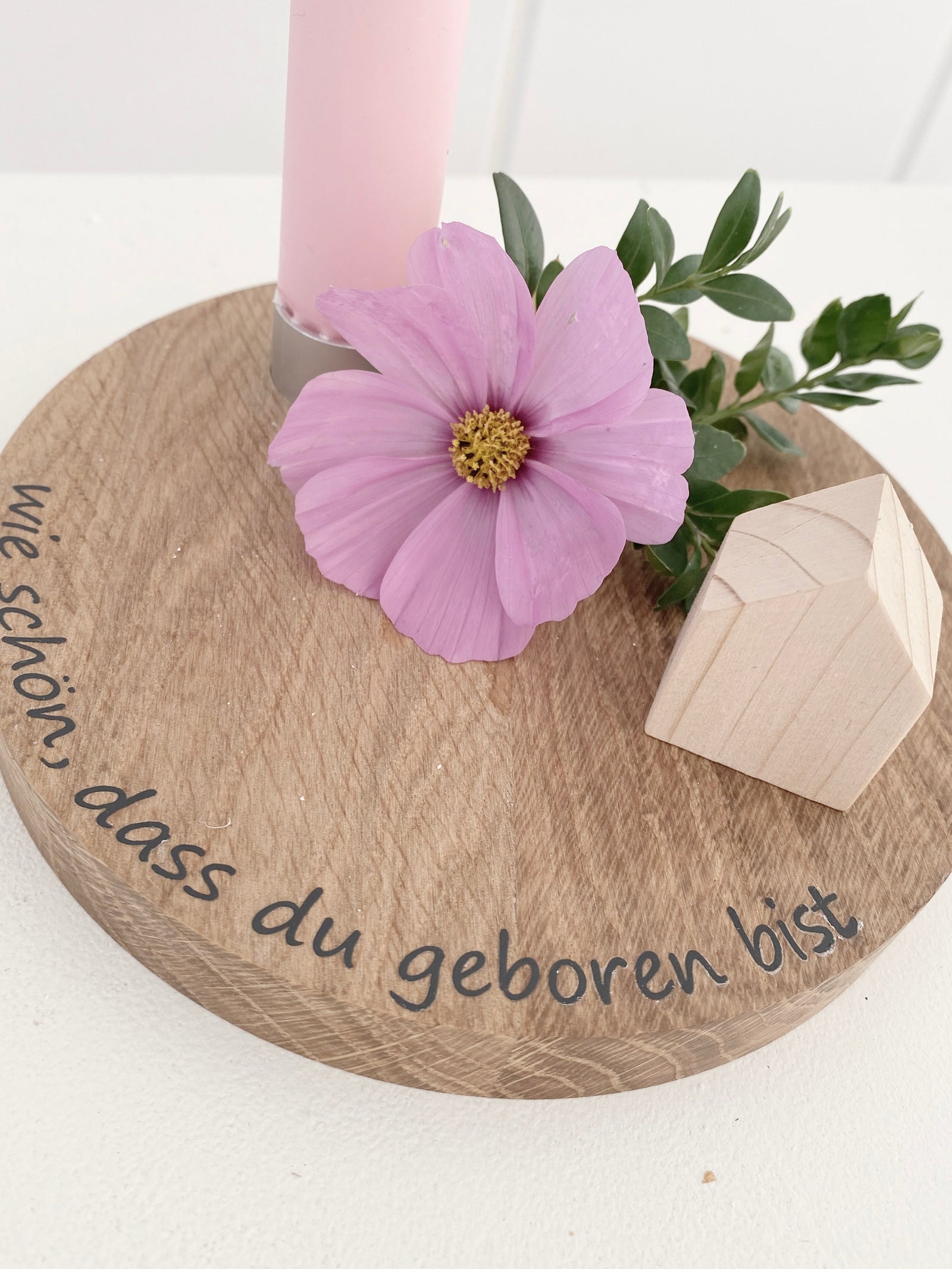 Rundes Brett aus Buche mit einem integrierten Kerzenhalter in dem eine pastellfarbene steht. Am Rand steht ein Schriftzug "wie schön, dass du geboren bist" (schwarze Plotterfolie), dekoriert mit einer rosafarbenen Kosmeeblüte und einem naturfarbenen Minihaus