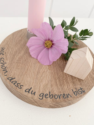 Detailaufnahme: rundes Brett aus Buche mit einem integrierten Kerzenhalter in dem eine pastellfarbene steht. Am Rand steht ein Schriftzug "wie schön, dass du geboren bist" (schwarze Plotterfolie), dekoriert mit einer rosafarbenen Kosmeeblüte und einem naturfarbenen Minihaus. Kann mit Namen individualisiert werden