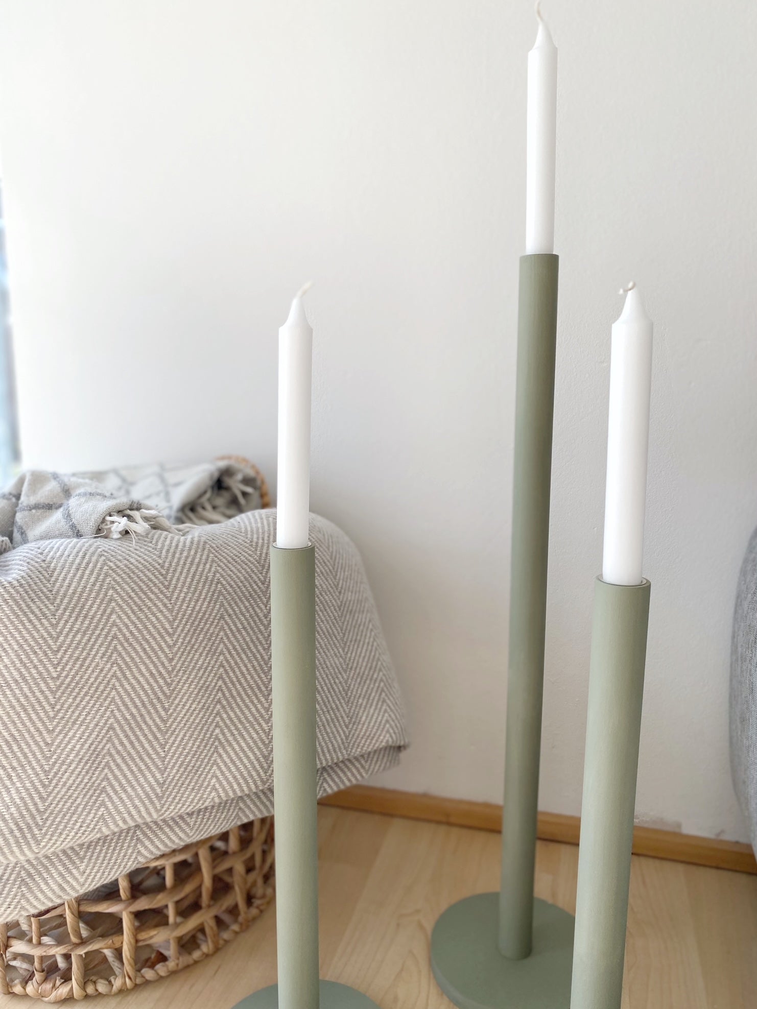 Drei olivfarbene große Kerzenständer mit weißen Kerzen, die neben dem Rand eines grauen Sofas stehen. Dahinter ein Jutekorb, in dem eine Wolldecke liegt.