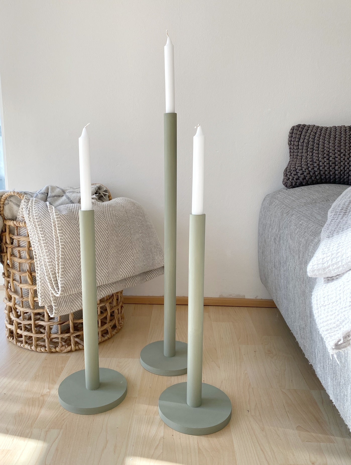 Drei olivfarbene große Kerzenständer mit weißen Kerzen, die neben dem Rand eines grauen Sofas stehen. Dahinter ein Jutekorb, in dem eine Wolldecke liegt.