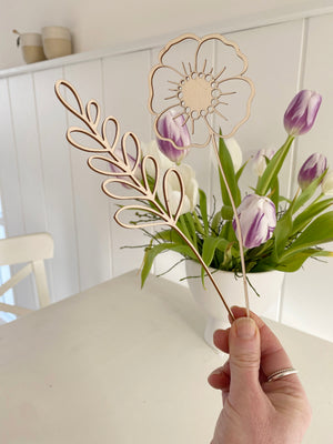 zwei lasercut Holzblumen in der Hand. Einmal wine minimalistische Blume, einmal ein Gras. Dahinter ein weißer Küchentisch mit einer Schale mit lila Tulpen und zwei Bechern