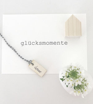 Mini Dekohaus auf einer Postkarte mit dem Aufdruck "glücksmomente". Darauf liegt ein kleiner Ahänger mit der Aufschrift "home", daneben liegen ein paar weiße Büte