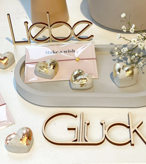 Hübsch dekoriert: ein ovales Beton tablett mt drei Betonherzen mit Blattgold, einem Armband und Schleierkraut. Dhinter steht ein Schriftzug "Liebe" aus Holz, davor ein Schriftzug, liegend "Glück"