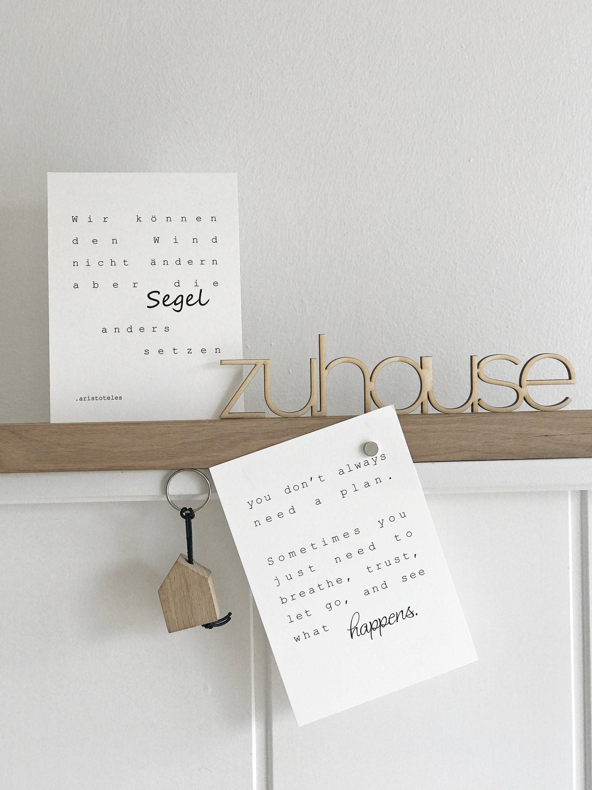 Holzleiste, dekoriert mit einer stehendes Postkarte, einer Postkarte die schräg daran mit einem Magneten befestigt ost. einem Schlüsselanhänger und einem Holz Schriftzug "zuhause"