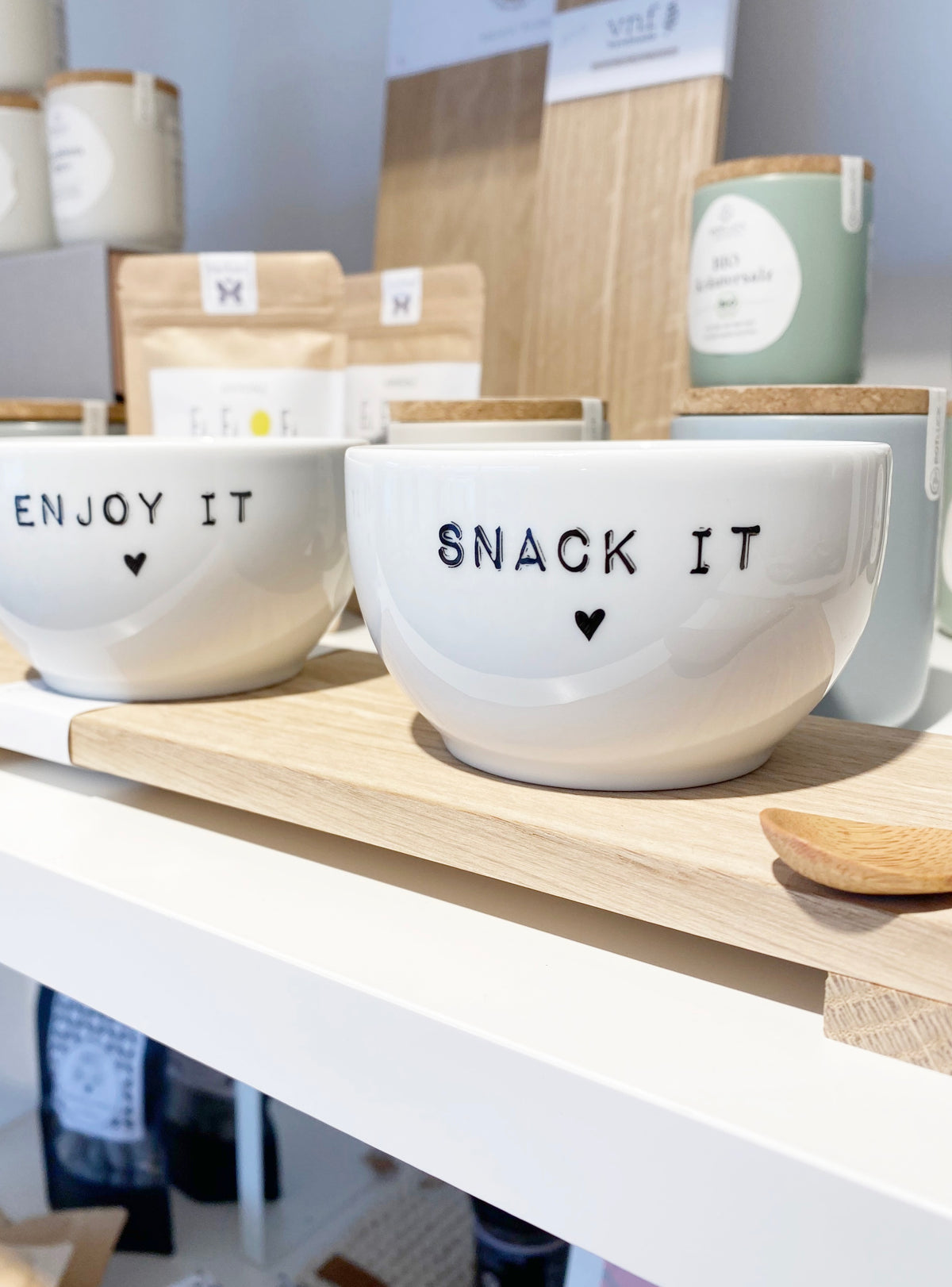 Eiblick in ein Regal im Conceptstore Stadtradstil: mehrer Küchenutensilien im Hintergrund, im Vordergrund zwei weiße, kleine Porzellan schalen mit den Aufdrucken "enjoy it" und "snack it"