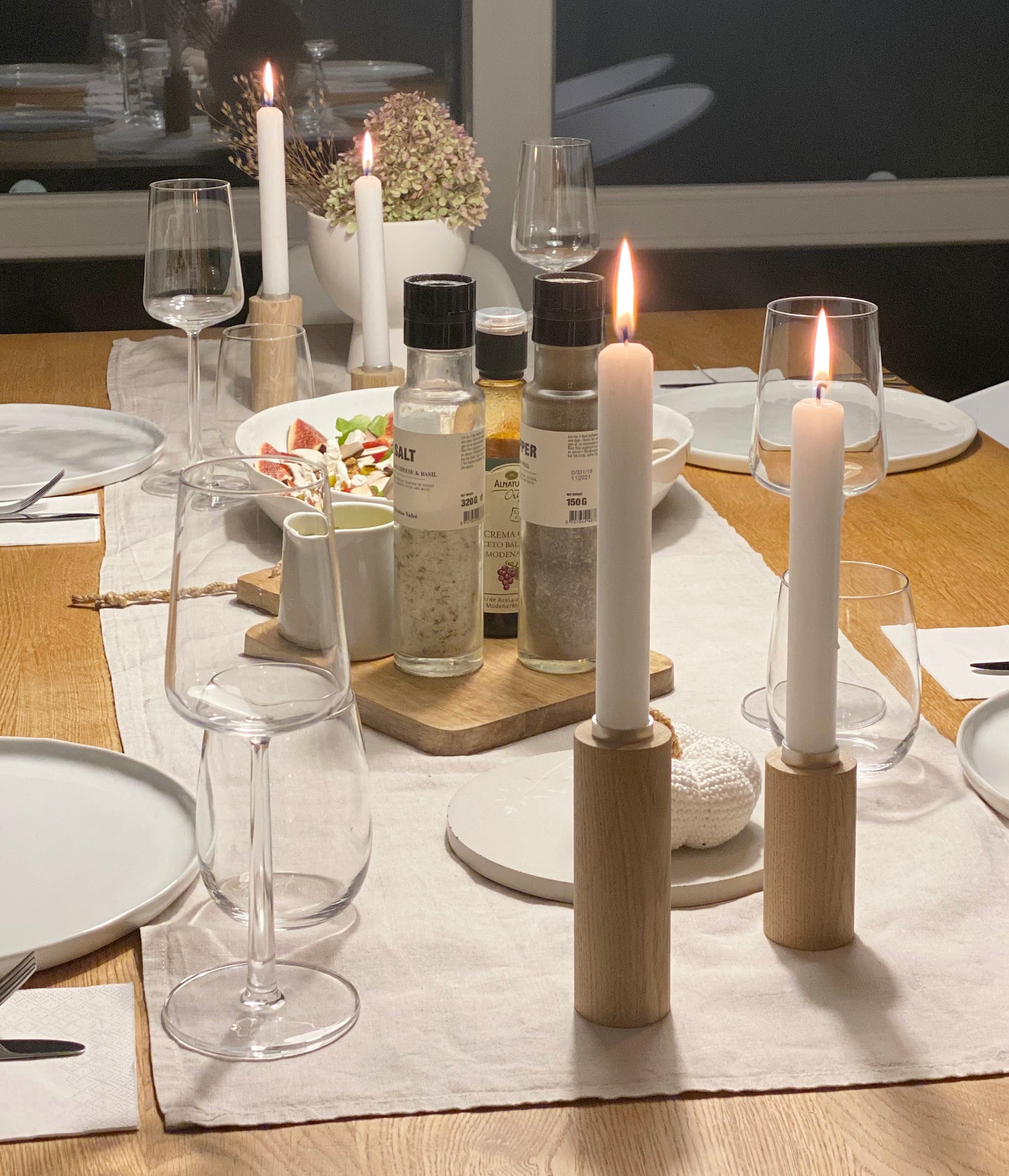 Eingedeckter Tisch,natürlich und unkompliziert mit Tischläufer auds Leinem verschieden groen Kerzenständern von Stadtrandstil mit weißen angezündeten Kerzen, weißen Tellern, Gläsern und einem Holzbrett, auf dem Salz, Pfeffer und Dressings stehen