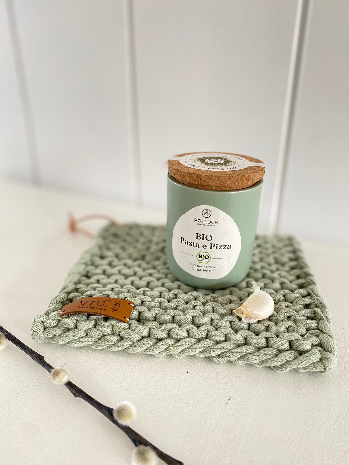 Salbeifarbener , handgestrickter Untersetzer, auf dem eine Knoblauchzehe liegt und ein farblcih passender Gewürztopf