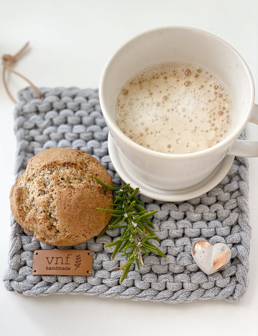 Aufsicht auf einen hellgrauen, handgetrickten Untersetzer mit Lederlabel. Darauf eine weiße Tasse mit Milchkaffe, einem kleinem Brötchen, einem Zweig Rosmarin und einem leinen Betonherz mit Blattgold