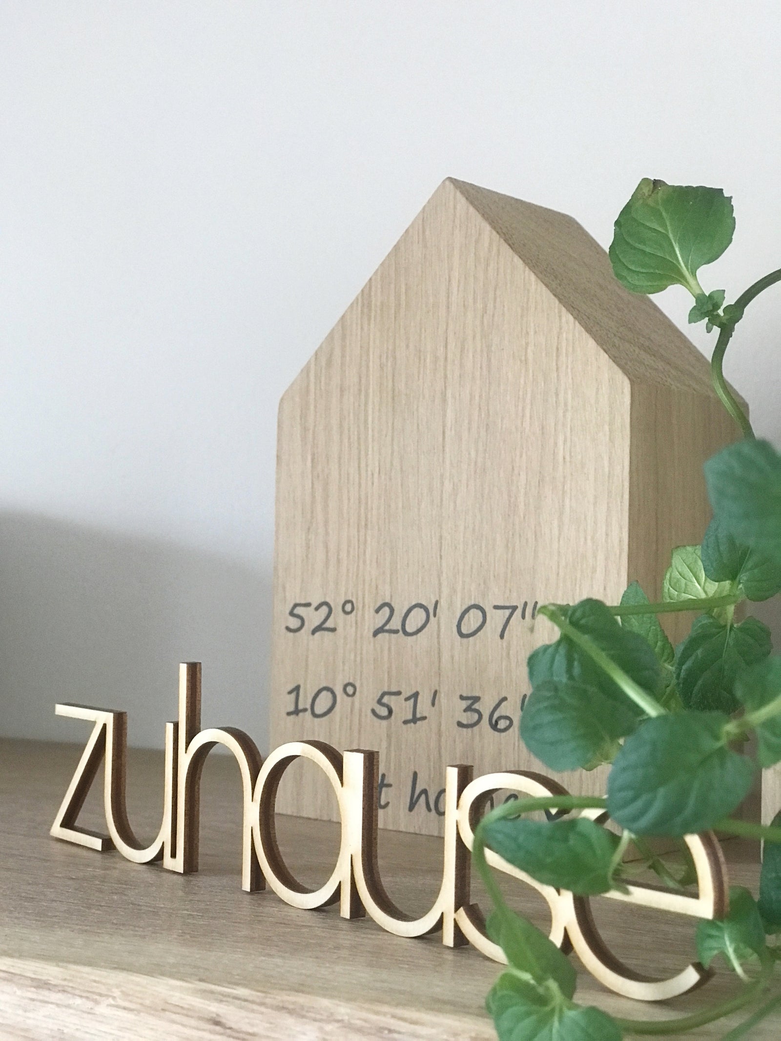 Schrägsicht auf ein Dekohaus aus Eichenholz, mit Koordinaten indiidualisiert, davor ein Schriftzug aus Holz "zuhause" unnd grüne Blätter.  Natürliche Dekoration
